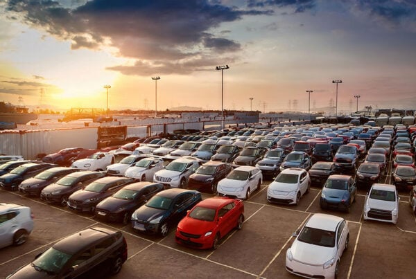 A large outdoor car auction site at sunset, with neatly lined rows of cars in various colours under tall floodlights, surrounded by industrial fencing and distant pylons against a warm evening sky.